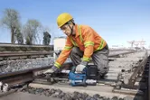 A worker wearing safety equipment uses a power tool to tighten bolts on railway tracks.