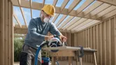 A person wearing safety equipment cuts wood with a cordless circular saw at a construction site.