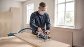 Person wearing safety equipment cuts wood with a cordless circular saw on a workbench.
