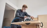 A person wearing safety equipment cuts wood with a cordless circular saw.