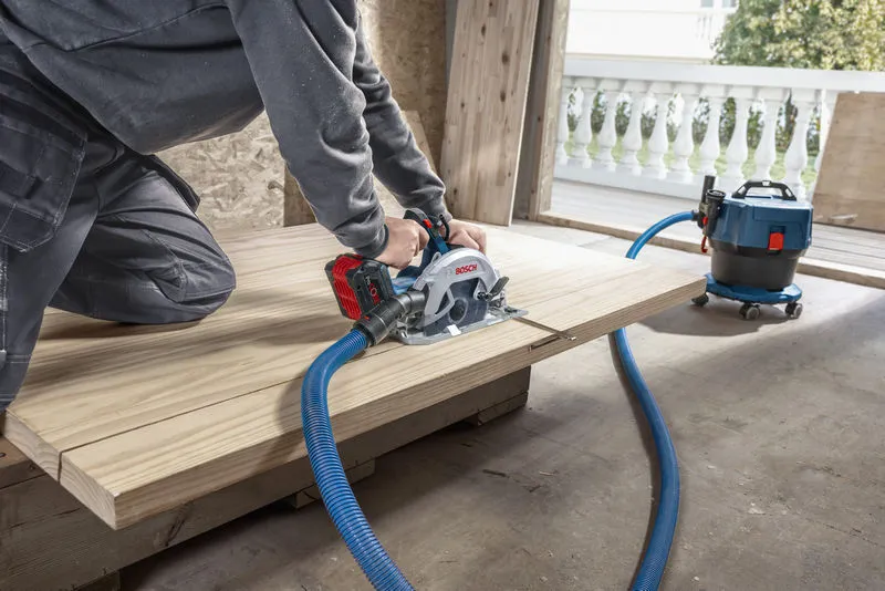 A person cuts wood with a cordless circular saw connected to a dust extractor.