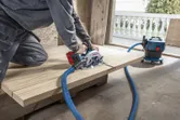 A person cuts wood with a cordless circular saw connected to a dust extractor.