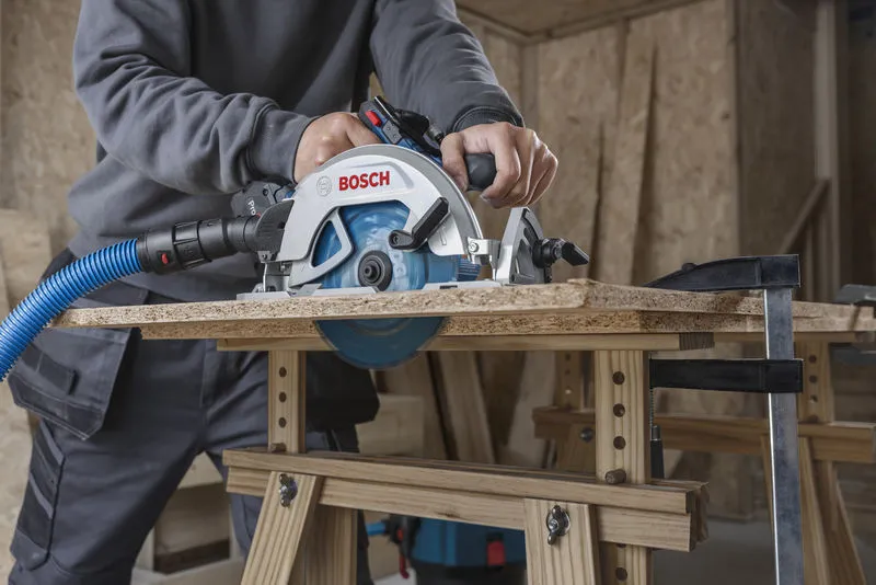 A person cuts wood with a cordless circular saw attached to a dust extraction hose.