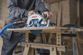 A person cuts wood with a cordless circular saw attached to a dust extraction hose.