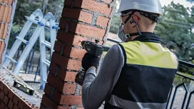 Person wearing safety equipment drilling into a brick column at a construction site.