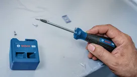 Person holding a screwdriver near a screw and a blue tool holder on a work surface.