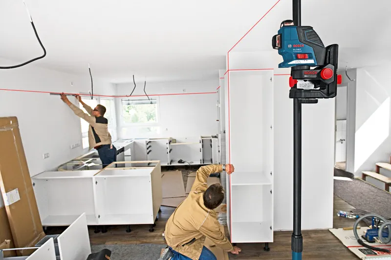 Two persons installing cabinets align shelves using a laser leveling tool.
