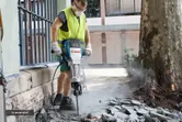 A person wearing safety equipment breaks pavement outdoors with a large electric breaker.