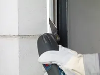 A worker wearing safety equipment cuts sealant from a window frame with a power tool.