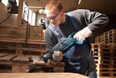 A person wearing safety equipment uses a reciprocating saw to cut a wooden pallet.