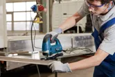 A person wearing safety equipment uses a jigsaw to cut a metal sheet.