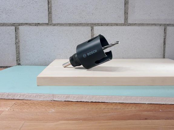 Hole saw drill bit placed on stacked wooden and drywall boards.