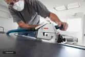 A person wearing safety equipment cuts a panel with a plunge saw attached to a guide rail.
