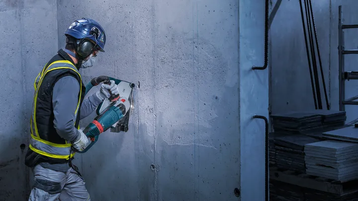 Εργαζόμενος που φοράει προστατευτικό εξοπλισμό κόβει έναν τσιμεντένιο τοίχο με ένα μεγάλο ηλεκτρικό εργαλείο.