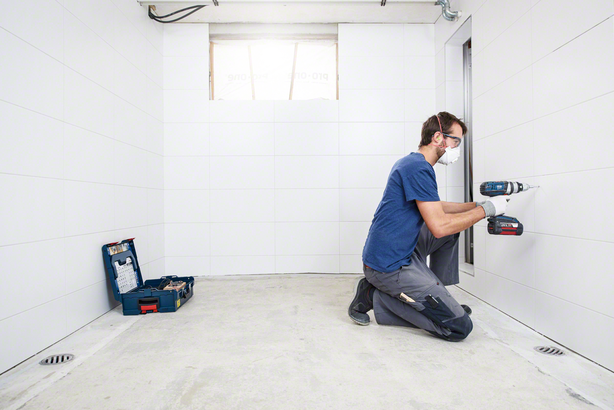 Osoba u zaštitnoj opremi buši popločani zid u praznoj sobi.