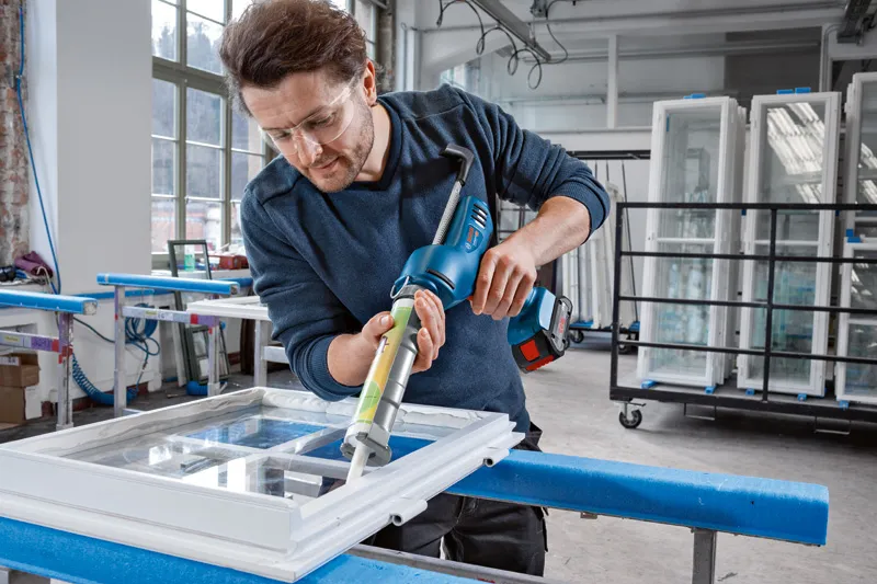 Osoba u zaštitnoj opremi nanosi brtvilo na okvir prozora bežičnim pištoljem za kit.