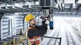Osoba koja nosi zaštitnu opremu upravlja magnetskom bušilicom na čeličnoj gredi iznad željezničkih tračnica.