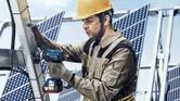 Osoba u zaštitnoj opremi zateže vijke na metalnom okviru u blizini solarnih panela.