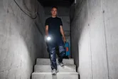 Osoba u zaštitnoj opremi hoda niz betonske stepenice držeći svjetiljku i kutiju s alatom.