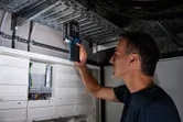Osoba pregledava metalni kabelski regal elektroničkim uređajem u blizini električnih panela.
