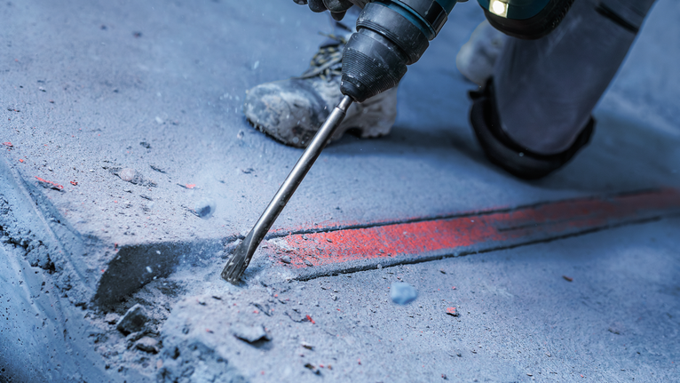 Osoba u zaštitnoj opremi dlijetom reže beton duž označene linije.