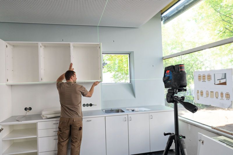 Osoba poravnava kuhinjske ormariće pomoću laserskog nivelatora na tronošcu.