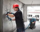 Egy védőfelszerelést viselő személy csiszol egy betonfalat, a közelben porelszívó működik.