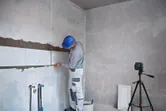 Egy biztonsági felszerelést viselő személy lézeres szintezővel csempéket rak le.