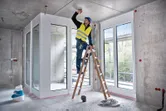 Egy biztonsági felszerelést viselő személy egy létrán áll, és a mennyezetbe fúr.