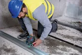 Biztonsági felszerelést viselő személy lézeres szintezővel igazítja a fémvázat a betonpadlón.