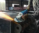 Egy védőfelszerelést viselő személy sarokcsiszolóval csiszolja az acélgerenda élét.