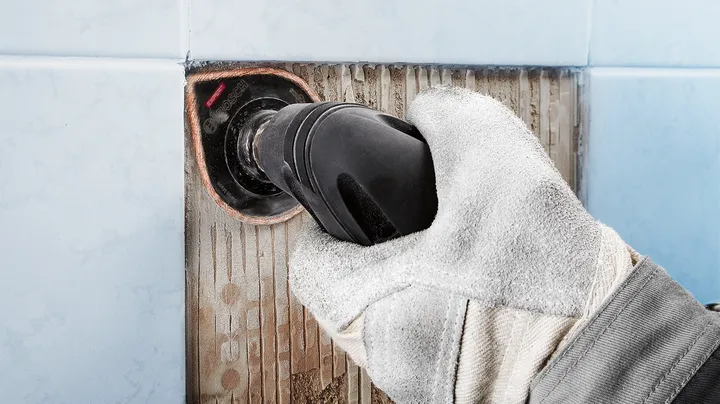 Biztonsági felszerelést viselő személy elektromos szerszámmal távolítja el a fugát a csempéről.