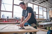 Egy biztonsági felszerelést viselő személy elektromos csiszolóval csiszol egy faajtót.