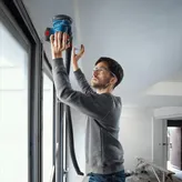 Egy biztonsági felszerelést viselő személy elektromos csiszolóval csiszolja a mennyezetet egy ablak közelében.
