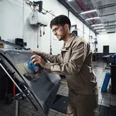 Egy biztonsági felszerelést viselő személy elektromos csiszolóval csiszolja az autó ajtaját.