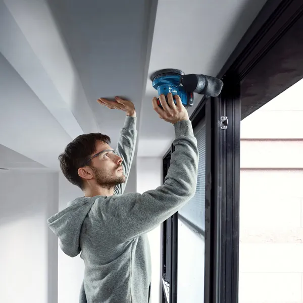 Egy biztonsági felszerelést viselő személy elektromos csiszolóval csiszolja a mennyezet szélét.