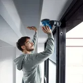 Egy biztonsági felszerelést viselő személy elektromos csiszolóval csiszolja a mennyezet szélét.