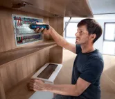 Egy személy akkus csavarhúzóval rögzíti az alkatrészeket egy elektromos panelben.