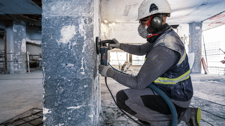 Biztonsági felszerelést viselő személy csiszol egy betonoszlopot egy építkezésen.