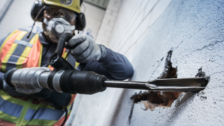 Biztonsági felszerelést viselő személy fúrókalapácsot használ egy betonfal áttöréséhez.
