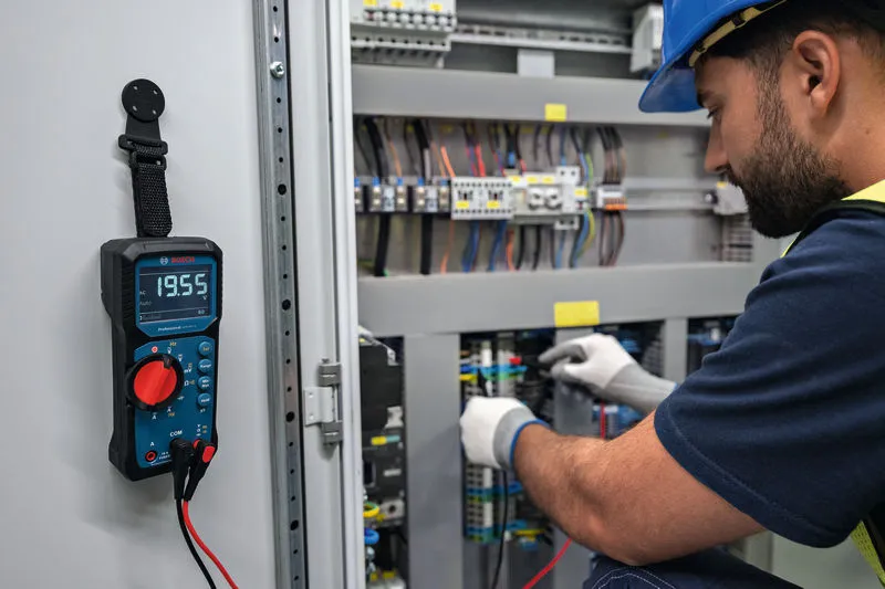 Biztonsági felszerelést viselő személy digitális multiméterrel teszteli az elektromos panelt.