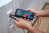 Egy személy digitális multiméterrel méri a feszültséget a fali aljzatban.