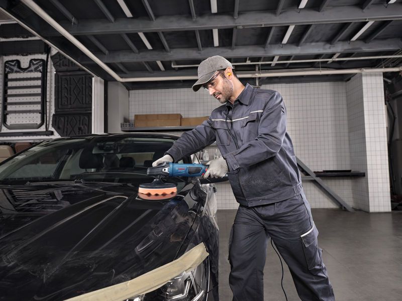 Egy biztonsági felszerelést viselő személy elektromos szerszámmal polírozza az autó motorháztetejét.