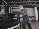 Egy biztonsági felszerelést viselő személy elektromos szerszámmal polírozza az autó motorháztetejét.