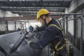 Egy biztonsági felszerelést viselő személy elektromos szerszámmal húzza meg az ipari csővezetékek csavarjait.