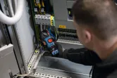 Egy biztonsági felszerelést viselő személy lakatmérővel ellenőrzi az elektromos panelt.