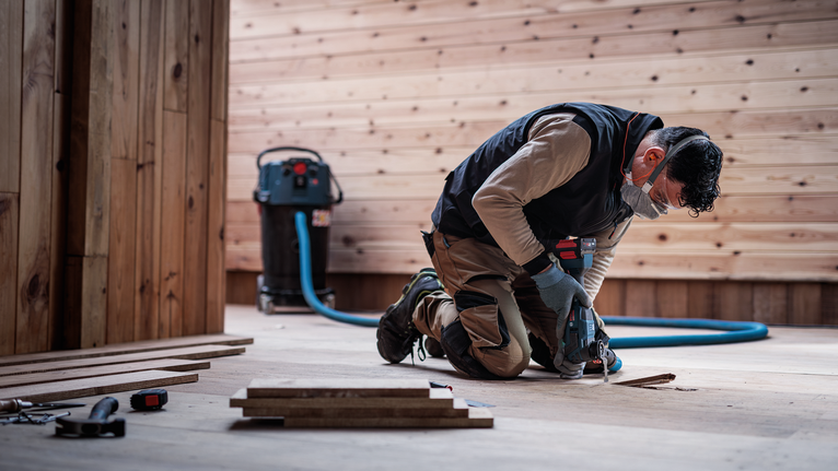 Egy biztonsági felszerelést viselő személy fúr egy fa padlót egy építkezési területen.