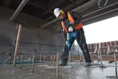 A person wearing safety equipment drills into concrete with a rotary hammer at a construction site.