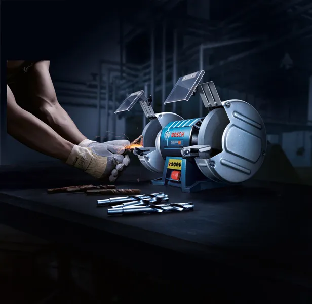 A person wearing safety equipment sharpens a drill bit with a double-wheeled bench grinder.