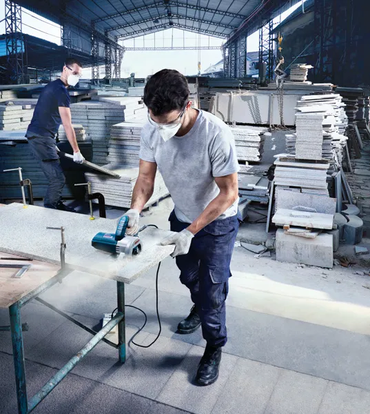 A person wearing safety equipment cuts a stone slab with a power saw in a warehouse.